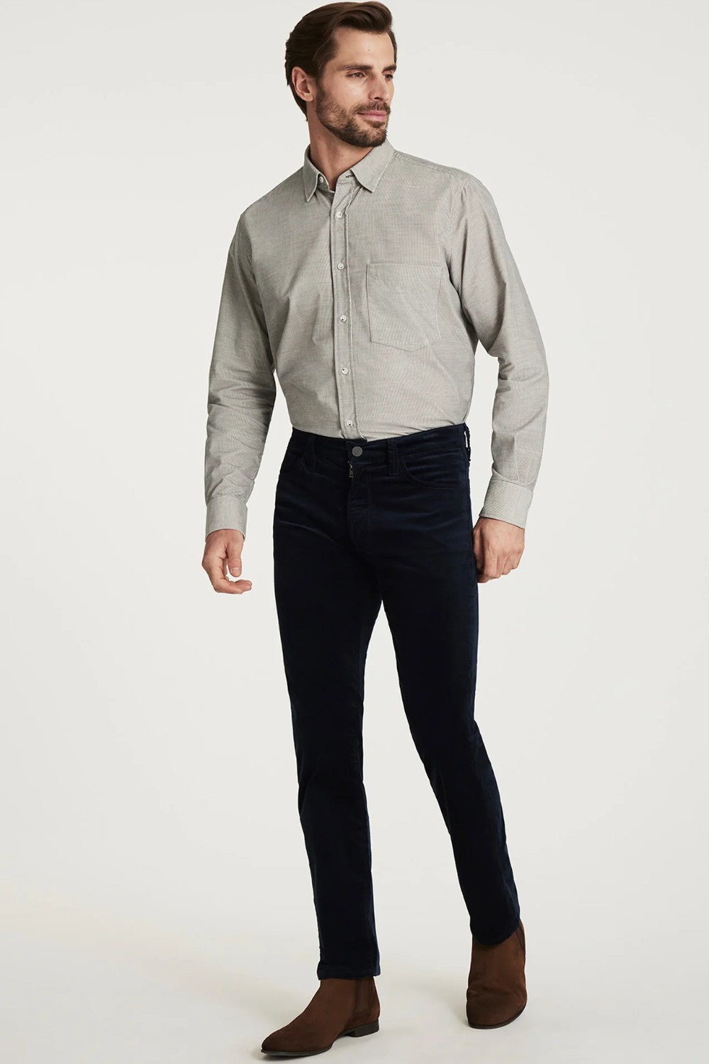 Man wearing a light gray button-up shirt, dark blue jeans, and brown shoes on a white background