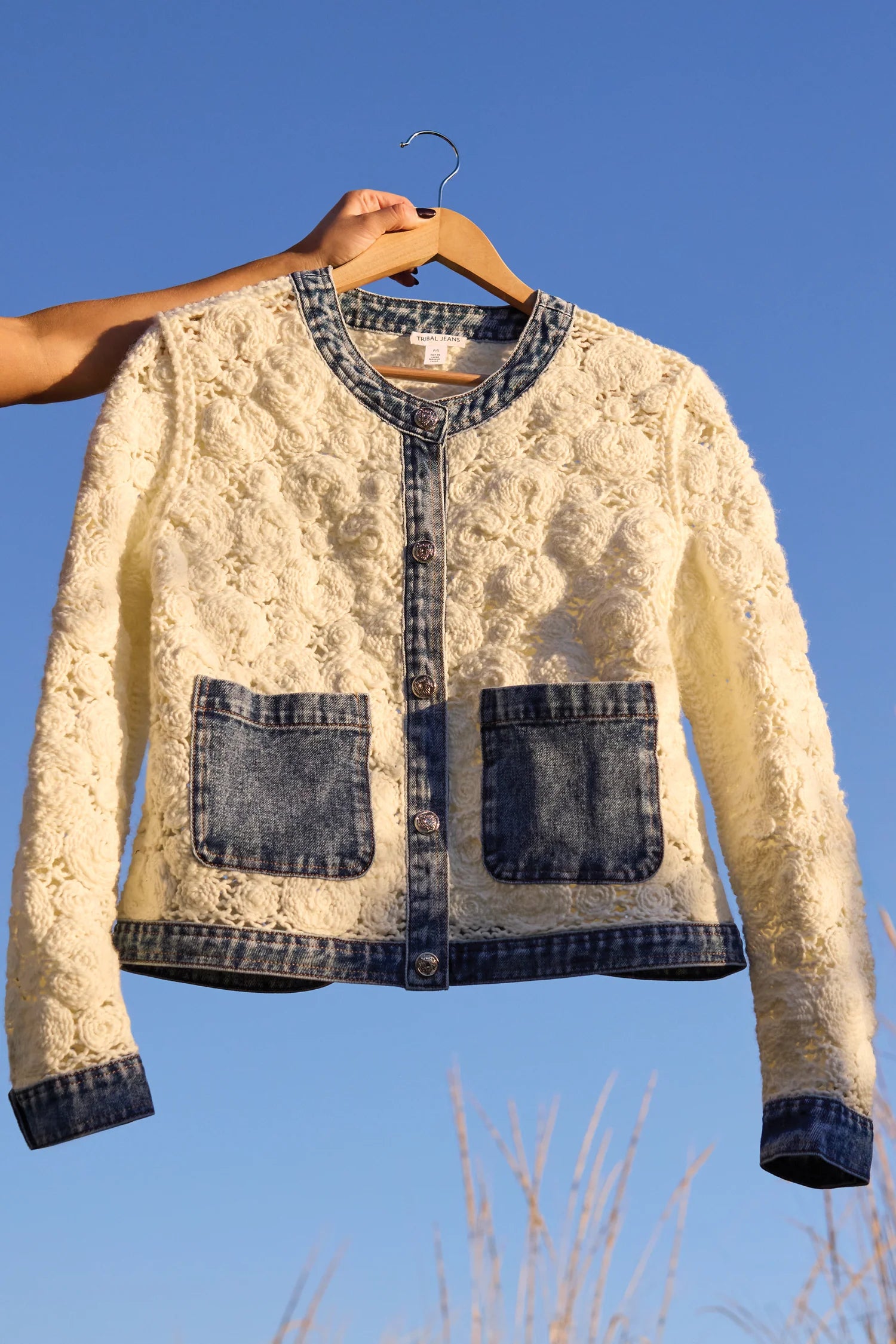White lace jacket with denim accents on a hanger against a blue sky.