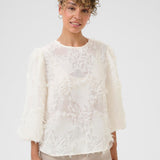 Woman wearing a white lace top and beige pants against a plain background