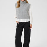 Woman wearing a gray sleeveless sweater over a white shirt with black pants on a white background