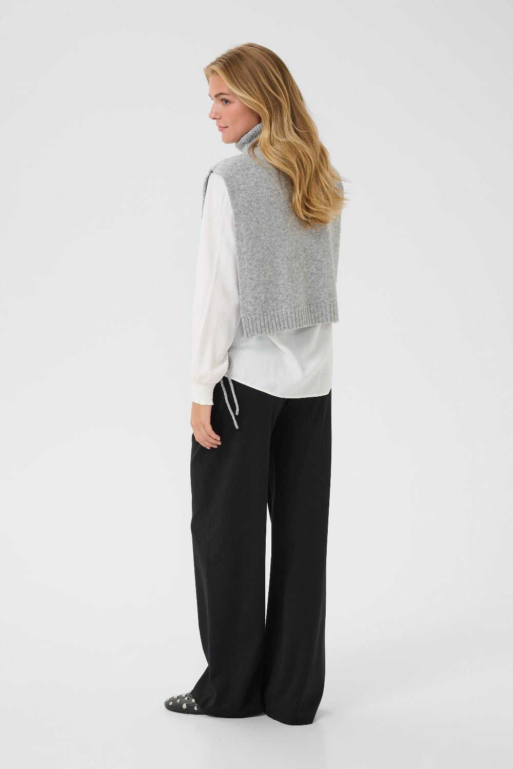Woman wearing a gray knitted vest over a white shirt with black pants on a white background