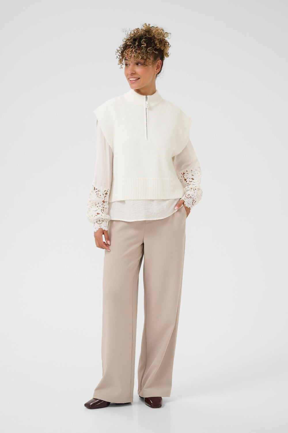 Woman wearing a white vest and blouse with lace sleeves and beige pants on a white background