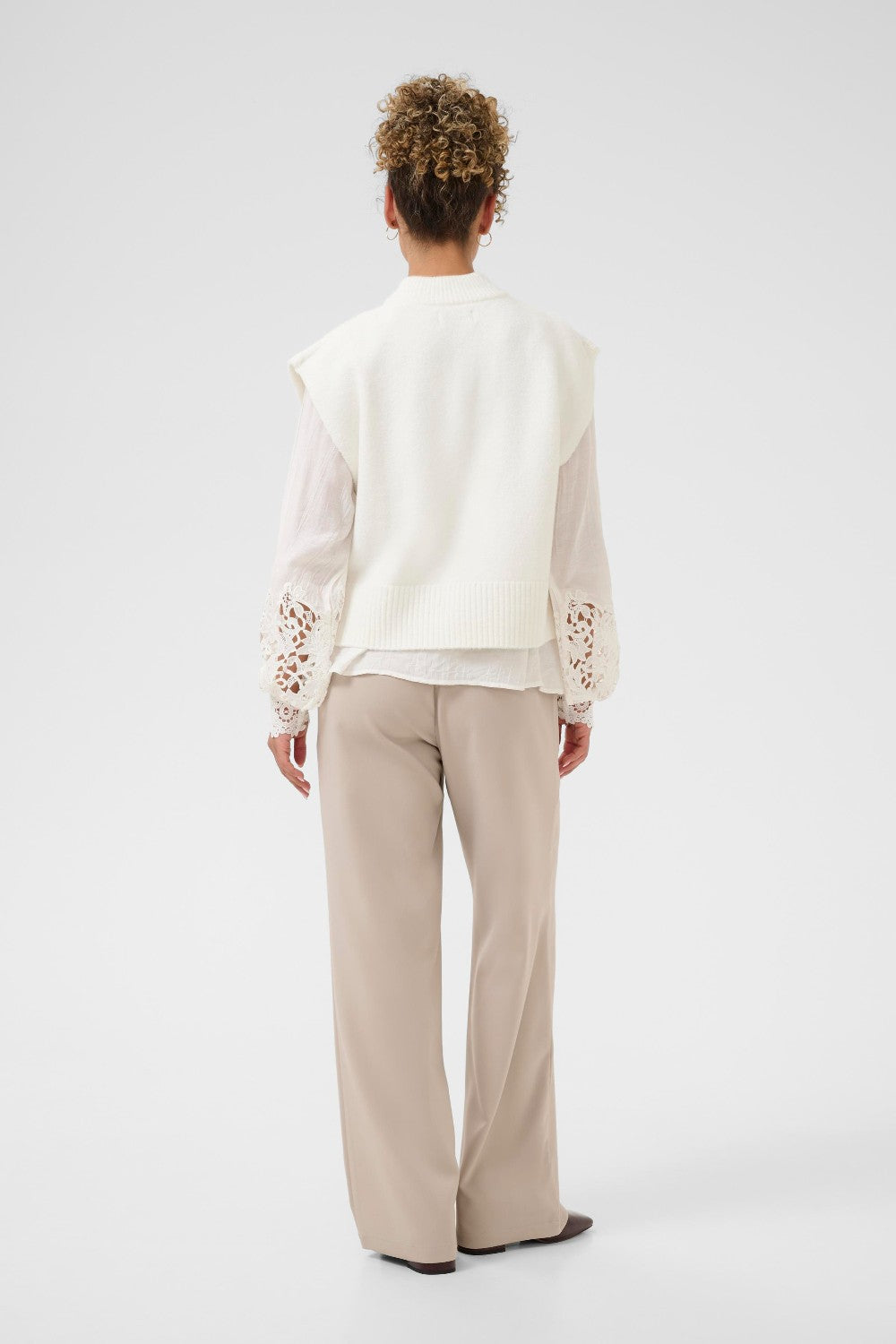 Person wearing a white vest and blouse with lace details and beige pants on a white background