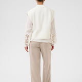 Person wearing a white vest and blouse with lace details and beige pants on a white background