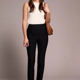 Woman wearing a white sleeveless top and black pants, holding a brown handbag against a plain background