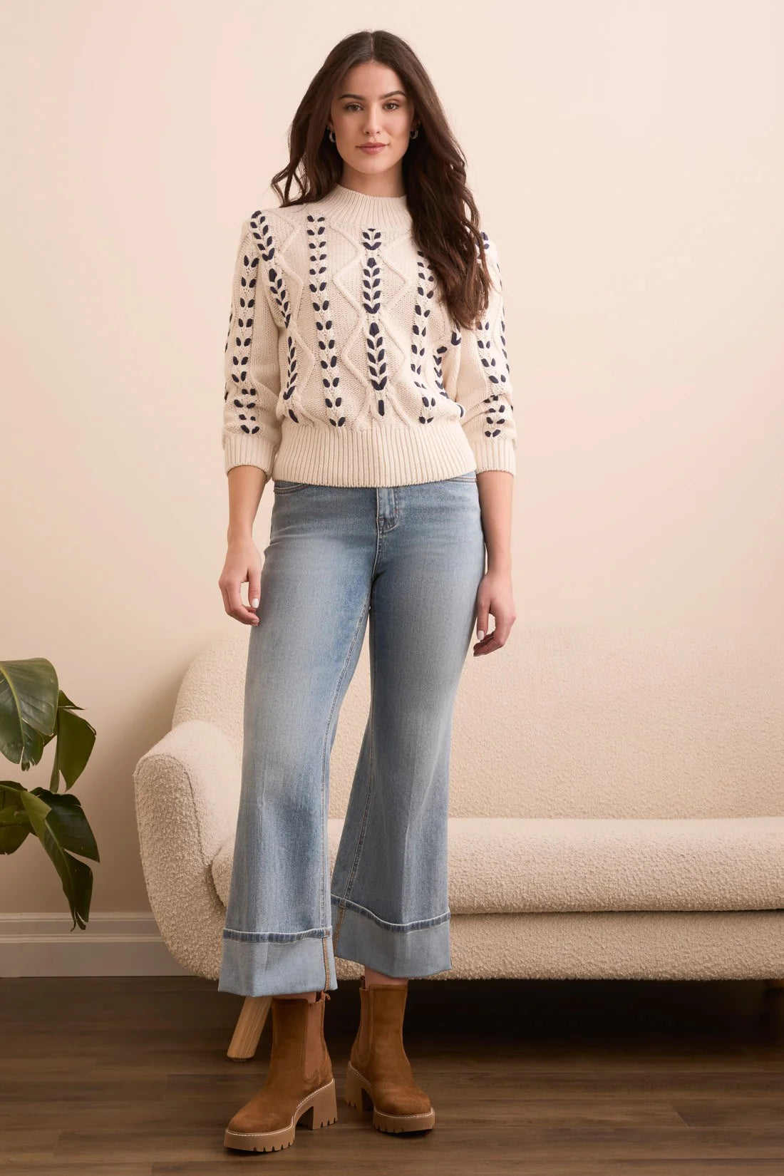 Woman wearing a cream sweater with black leaf patterns, light blue jeans, and brown boots in a room with a beige sofa and plant.