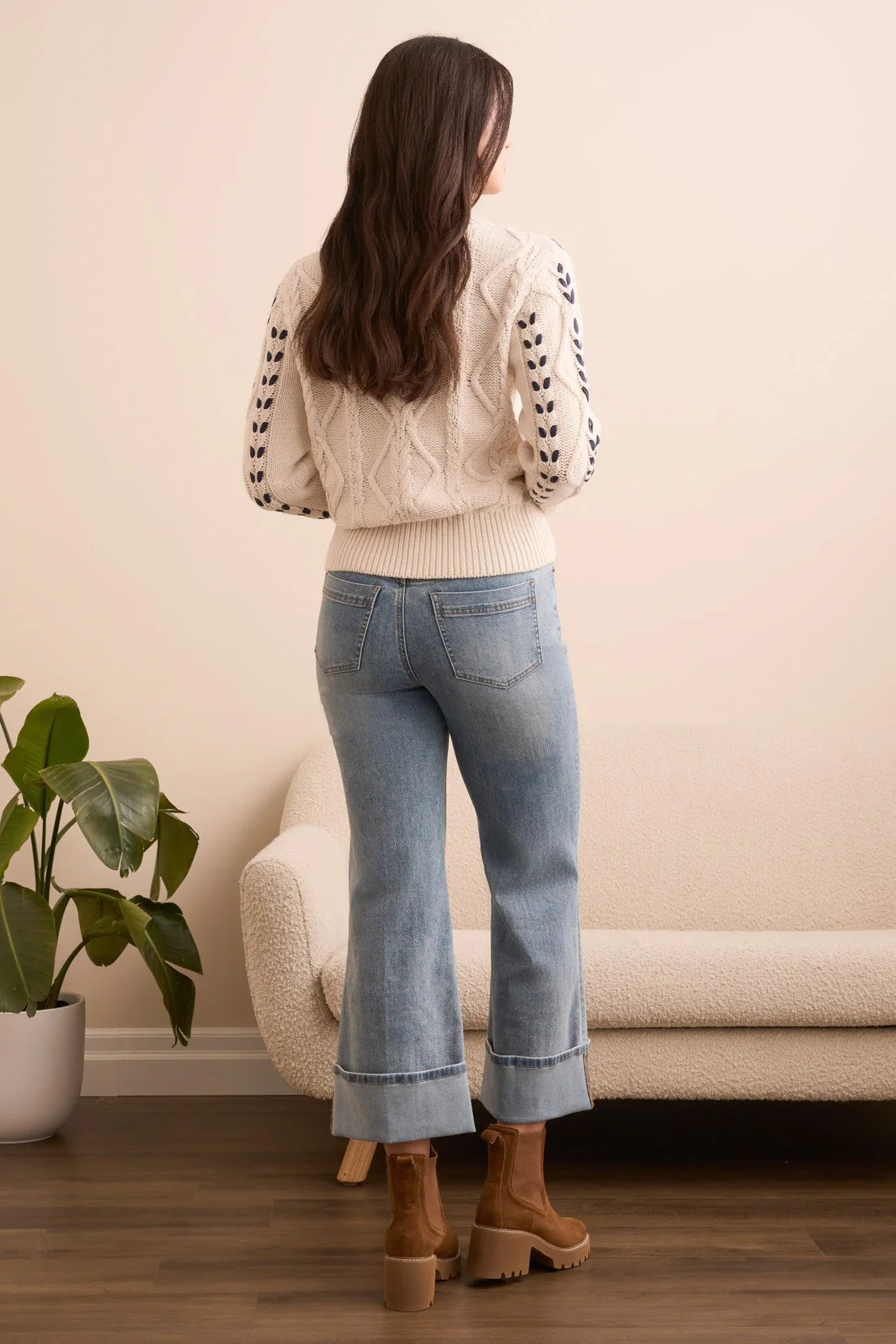 Woman wearing a white cable knit sweater and blue jeans standing in a room with a plant and sofa.
