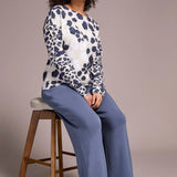 Woman sitting on a stool wearing a patterned top and blue pants against a plain background