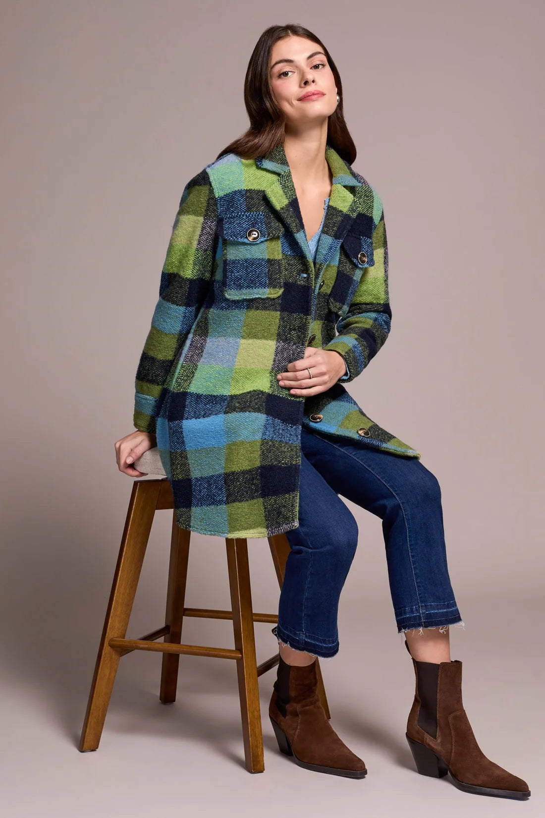 Woman wearing a plaid coat sitting on a wooden stool against a plain background