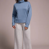 Woman wearing a blue sweater and light gray pants against a plain background