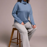 Woman wearing a blue turtleneck sweater and gray pants sitting on a wooden stool against a plain background