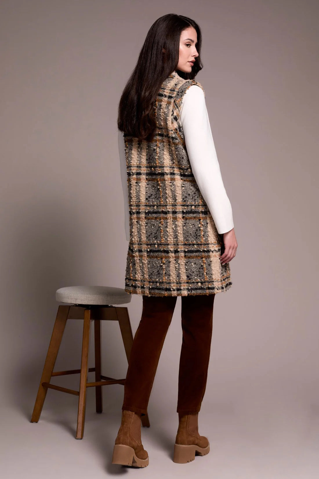 Woman wearing a plaid dress with a white shirt underneath, standing next to a wooden stool against a plain background.