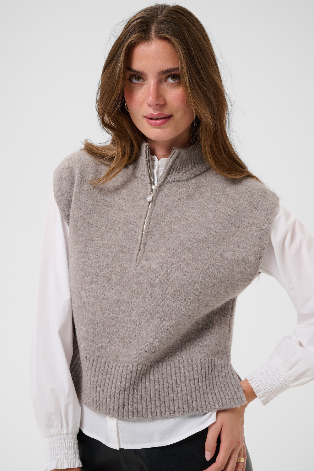 Woman modelling a brown half zip knit vest with a white shirt and black pants against a light backdrop.