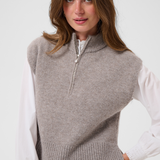 Woman modelling a brown half zip knit vest with a white shirt and black pants against a light backdrop.
