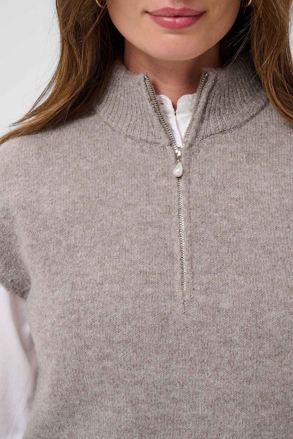 Close up of a woman modelling a brown half zip knit vest with a white shirt against a light backdrop.