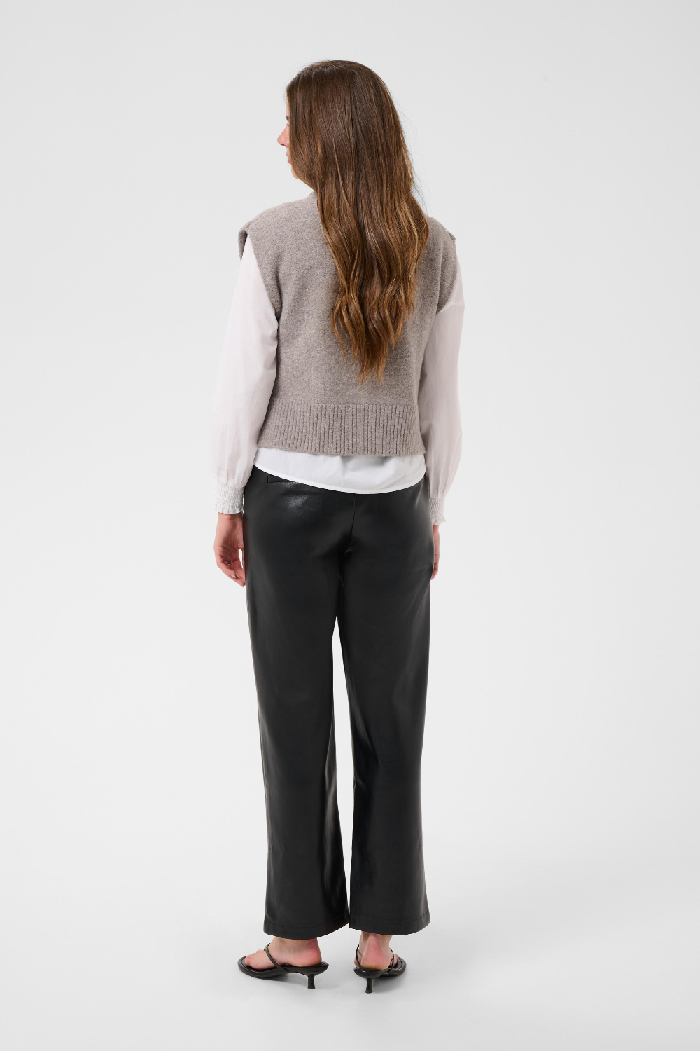 Woman, shown from the back, modelling a brown half zip knit vest with a white shirt and black pants black kitten heels against a light backdrop.