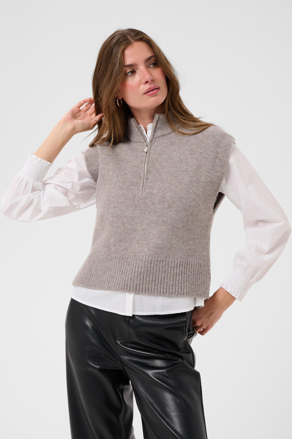Woman modelling a brown half zip knit vest with a white shirt and black pants against a light backdrop.