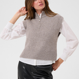 Woman modelling a brown half zip knit vest with a white shirt and black pants against a light backdrop.