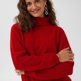Woman modelling a red turtle neck sweater with black pants against a light backdrop.