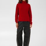 Woman modelling a red turtle neck sweater with black pants and black boots against a light backdrop.