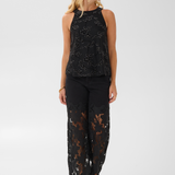 Woman modelling a sleeveless embellished black top and black pants  with black heels photographed against a light background.