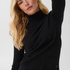 Woman modelling a black turtleneck with black pleather pants against a light backdrop.