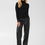 Woman modelling a black turtleneck with black pleather pants and black boots against a light backdrop.