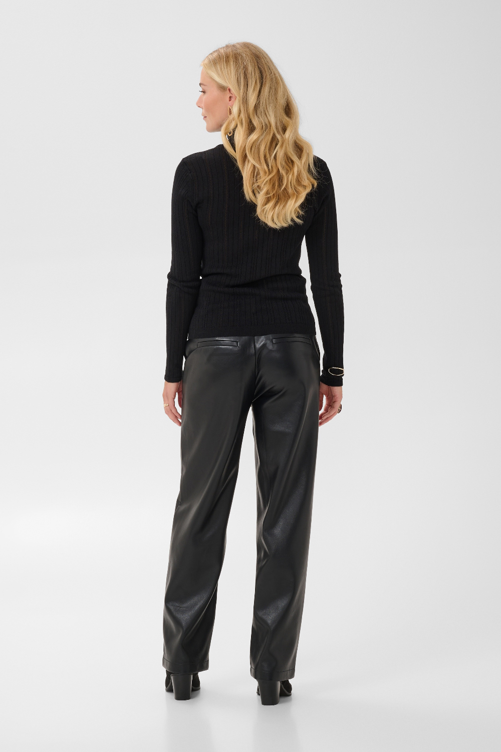 Woman, shown from behind, modelling a black turtleneck with black pleather pants and black boots against a light backdrop.