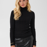 Woman modelling a black turtleneck with black pleather pants against a light backdrop.