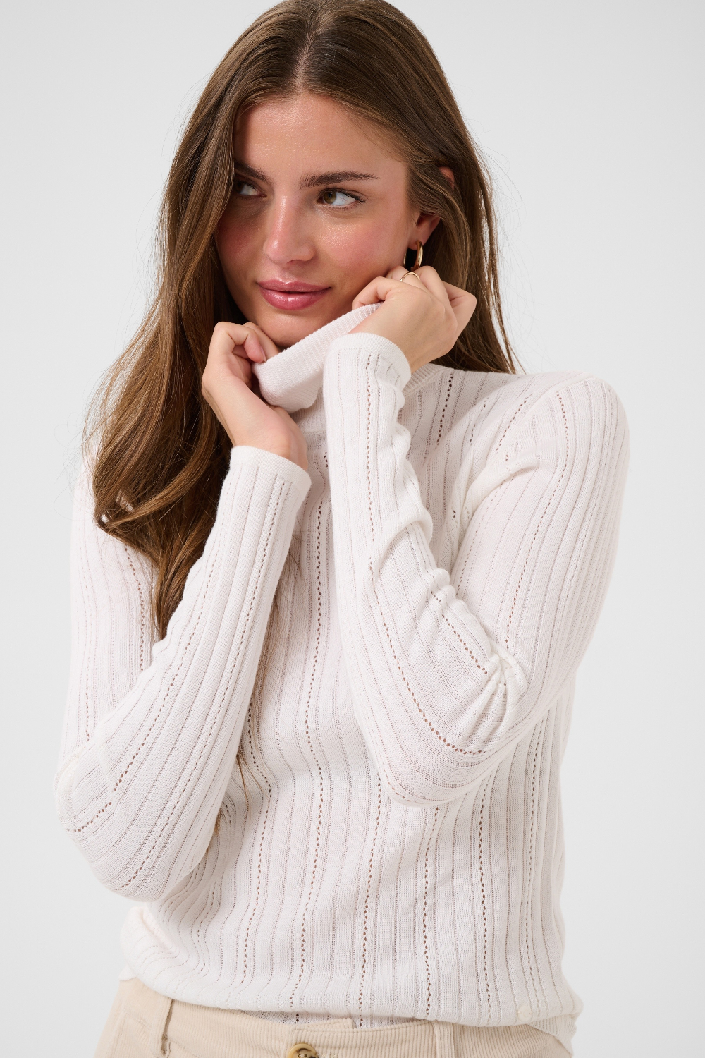 Woman modelling a cream turtleneck with beige corduroy pants against a light backdrop.