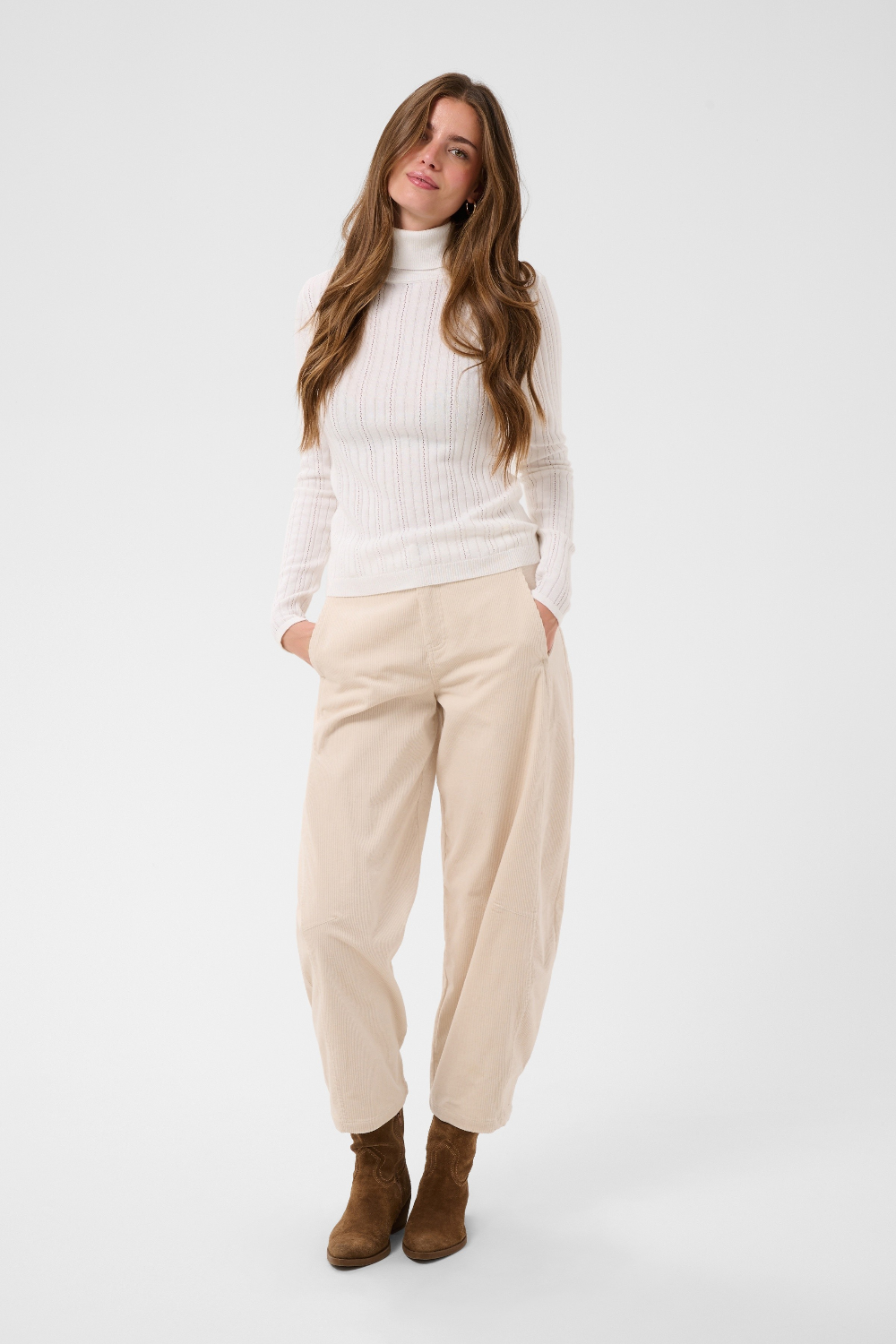 Woman modelling a cream turtleneck with beige corduroy pants and brown boots against a light backdrop.