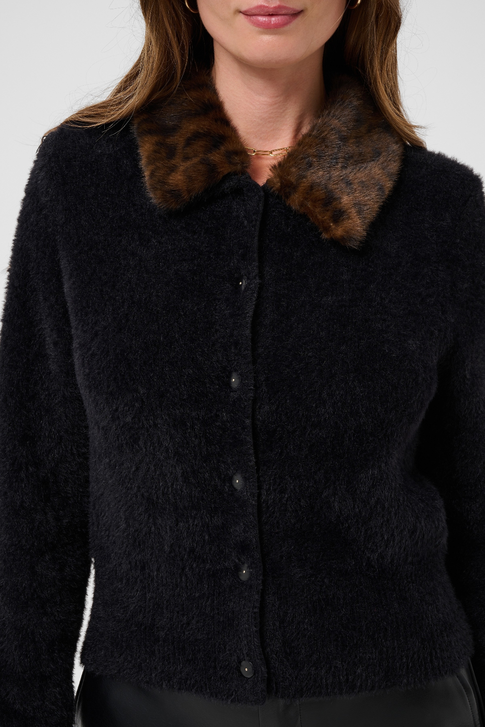 Close up of a woman modelling a black button front cardigan, with an animal print collar, with black pants, photographed against a light backdrop.