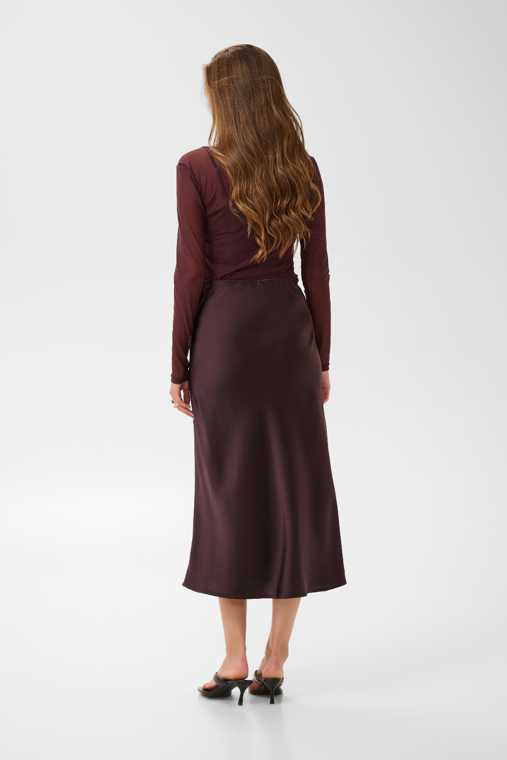 Woman, shown from the back, modelling a deep wine coloured sateen skirt with a mesh top and black kitten heels photographed against a light background.