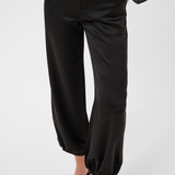 Woman modelling a sateen black pant with kitten heels and a black blouse photographed against a light backdrop.