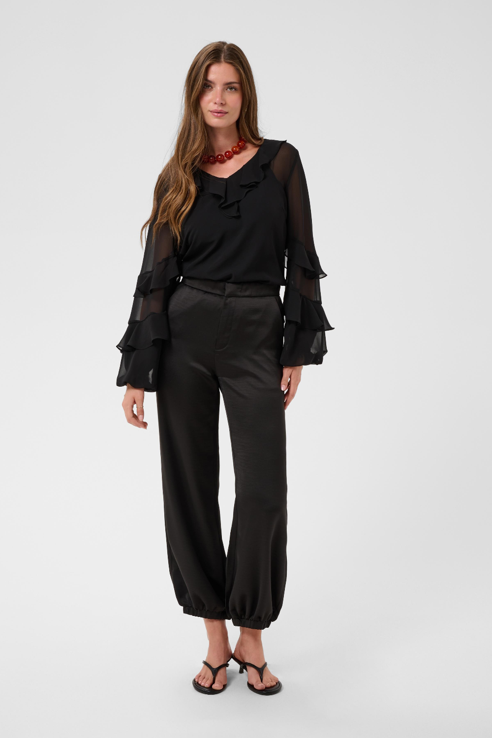 Woman modelling a sateen black pant with kitten heels and a black blouse photographed against a light backdrop.