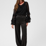Woman modelling a sateen black pant with kitten heels and a black blouse photographed against a light backdrop.