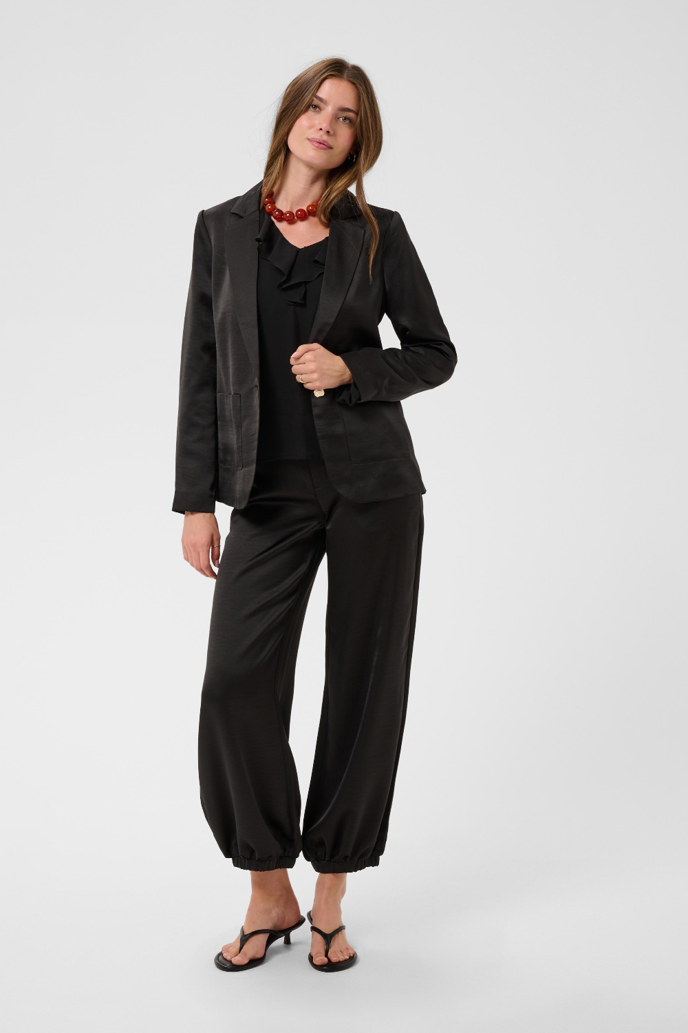 Woman modelling a black sateen blazer, black sateen pants with kitten heels and a black blouse photographed against a light backdrop.