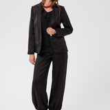 Woman modelling a black sateen blazer, black sateen pants with kitten heels and a black blouse photographed against a light backdrop.