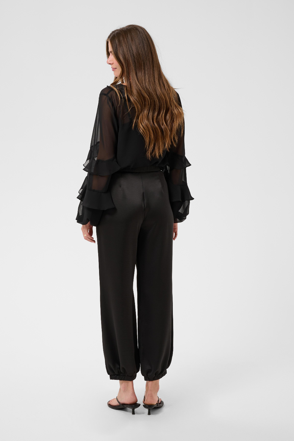 Woman, shown from behind, modelling a sateen black pant with kitten heels and a black blouse photographed against a light backdrop.
