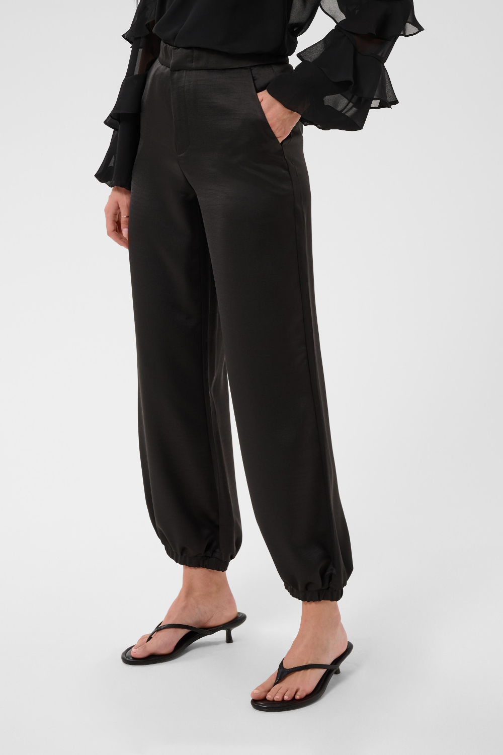 Woman modelling a sateen black pant with kitten heels and a black blouse photographed against a light backdrop.