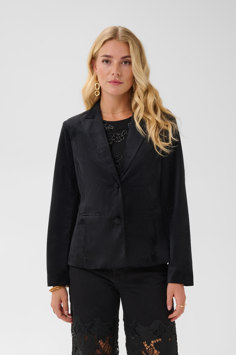 Woman modelling a black velour blazer with a sparkly black top and black pants photographed on a light background.