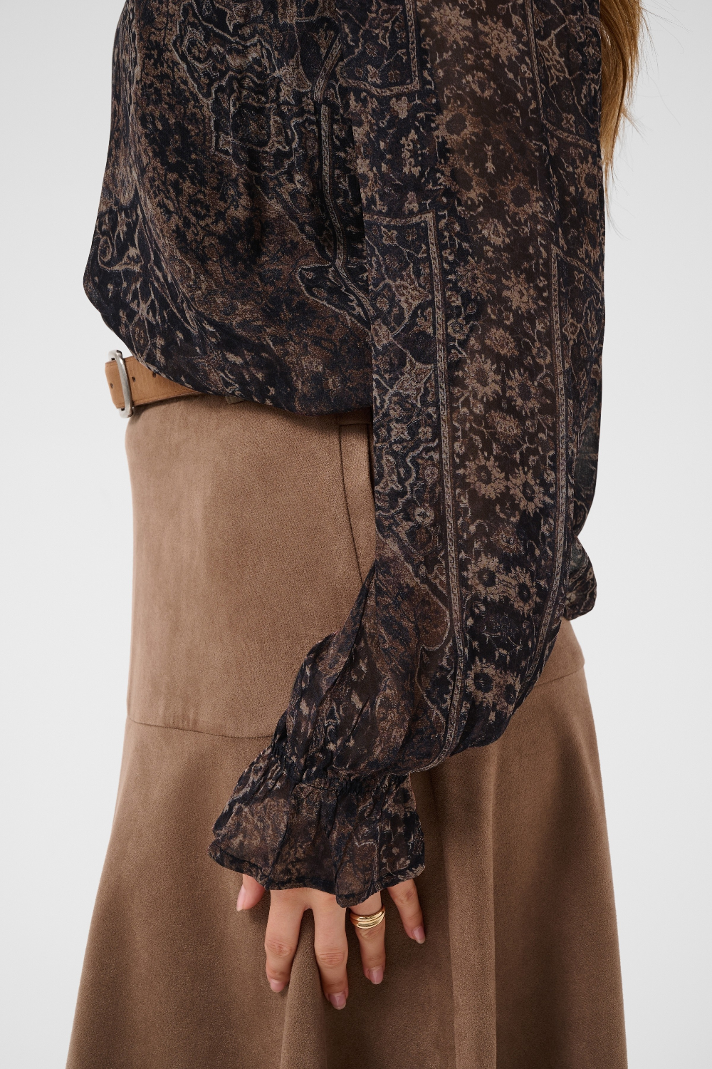 Close up of a woman modelling a printed brown v neck blouse with a tan skirt and belt photographed against a light background.
