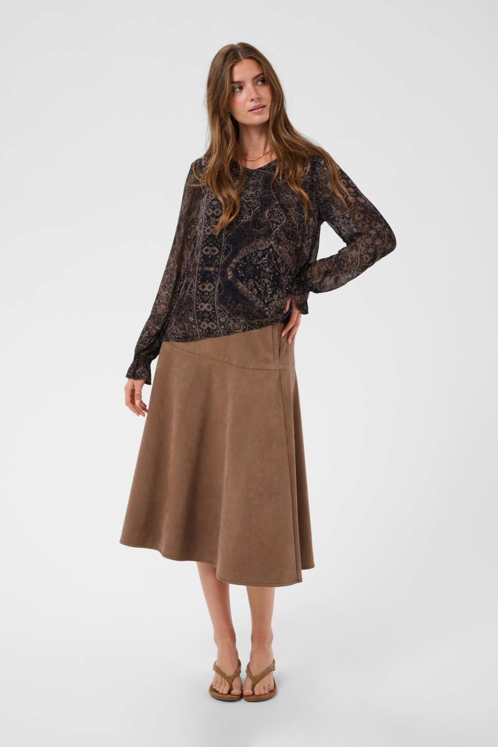 Woman modelling a printed brown v neck blouse with a tan skirt and shoes photographed against a light background.