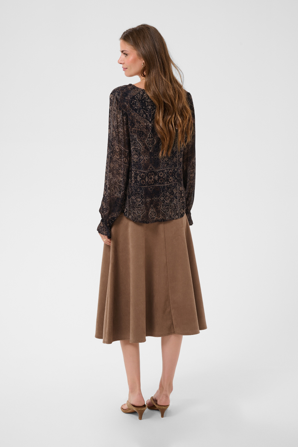 Woman modelling a printed brown v neck blouse with a tan skirt and shoes photographed against a light background.