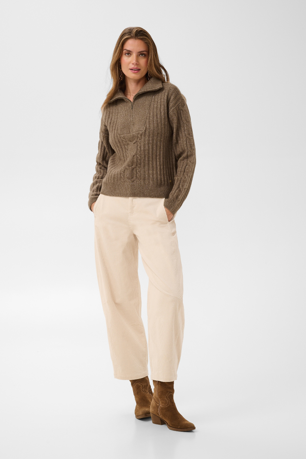 Woman modelling a brown 1/4 zip sweater with cream pants and brown boots photographed against a light backdrop.