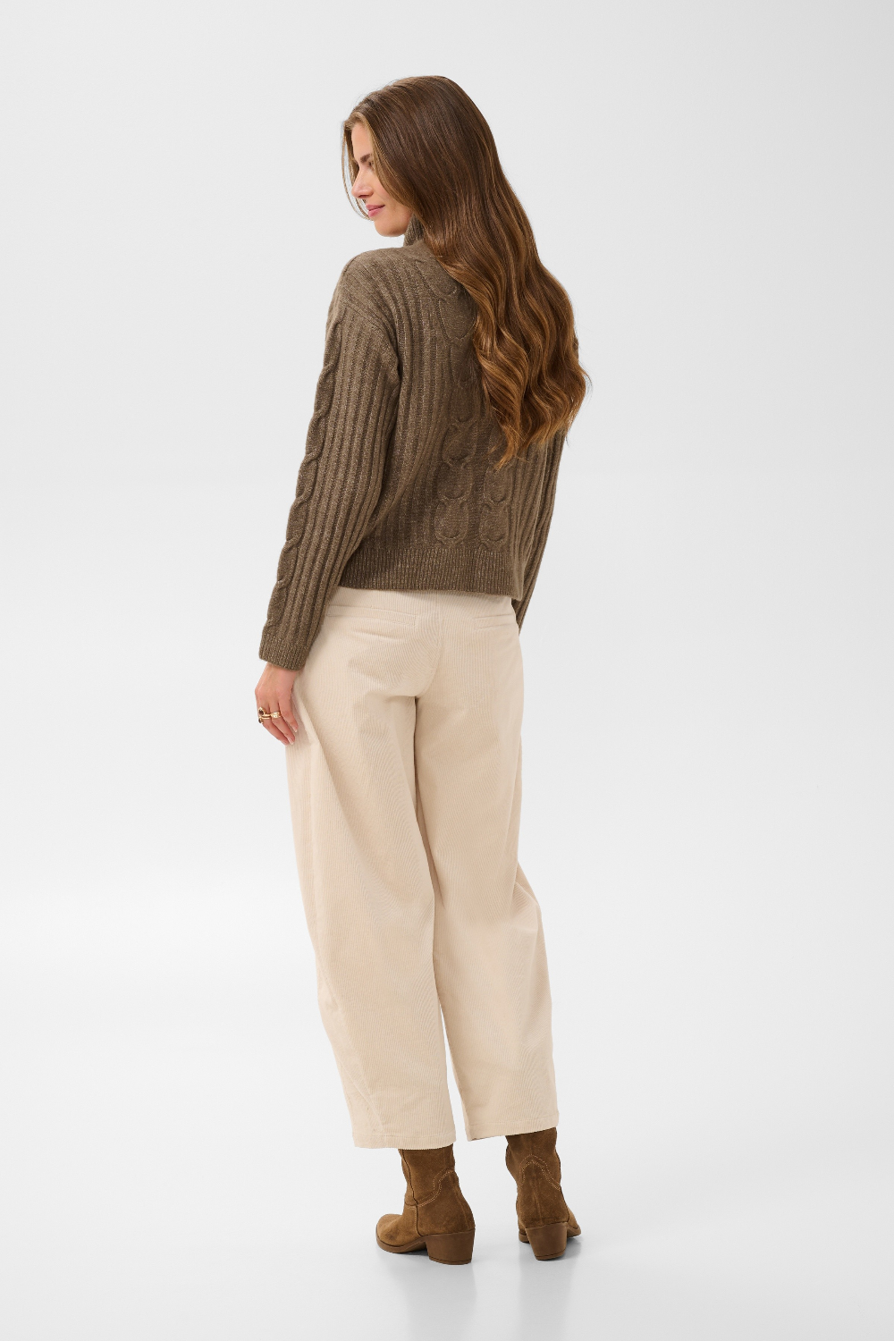 Woman, shown from behind, modelling a brown sweater with cream pants and brown boots photographed against a light backdrop.