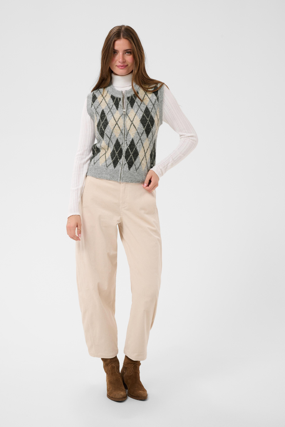 A woman modeling a sleeveless argyle knit vest over a white long-sleeve turtleneck, paired with cream trousers and brown ankle boots, against a light gray studio background.
