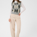 A woman modeling a sleeveless argyle knit vest over a white long-sleeve turtleneck, paired with cream trousers and brown ankle boots, against a light gray studio background.