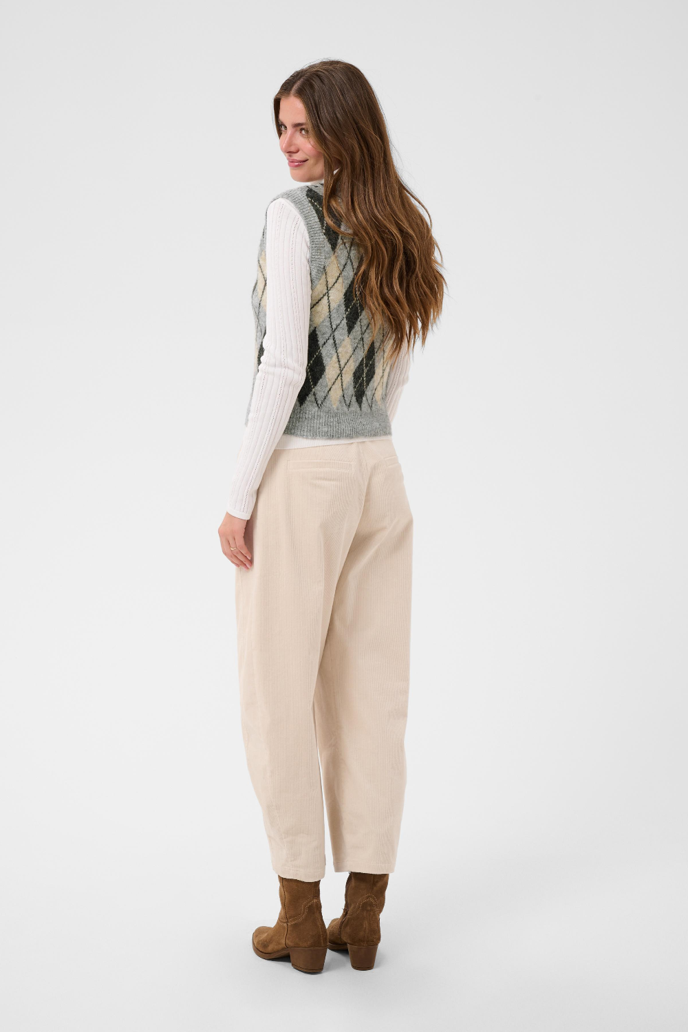 A woman, shown from behind, modeling a sleeveless argyle knit vest over a white long-sleeve turtleneck, paired with cream trousers and brown ankle boots, against a light gray studio background.