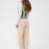 A woman, shown from behind, modeling a sleeveless argyle knit vest over a white long-sleeve turtleneck, paired with cream trousers and brown ankle boots, against a light gray studio background.